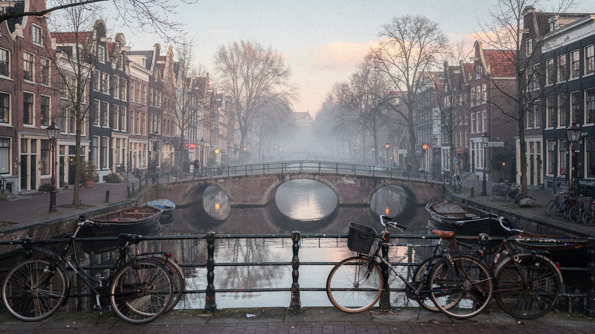 Gabled canal houses reflected in the still water of the Reguliersgracht at dawn, with a row of arched brick bridges receding into soft mist