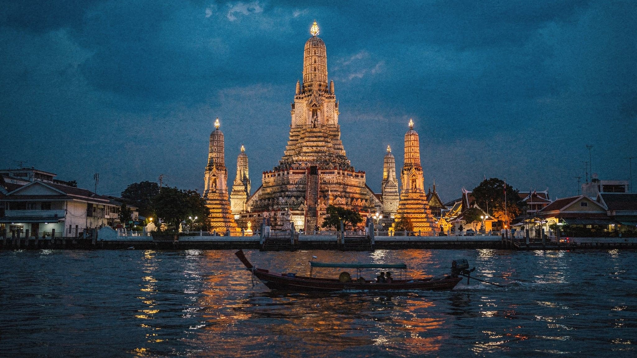 Wat Arun's central spire lit at blue hour across the Chao Phraya River, with a long-tail boat crossing the water in front of it