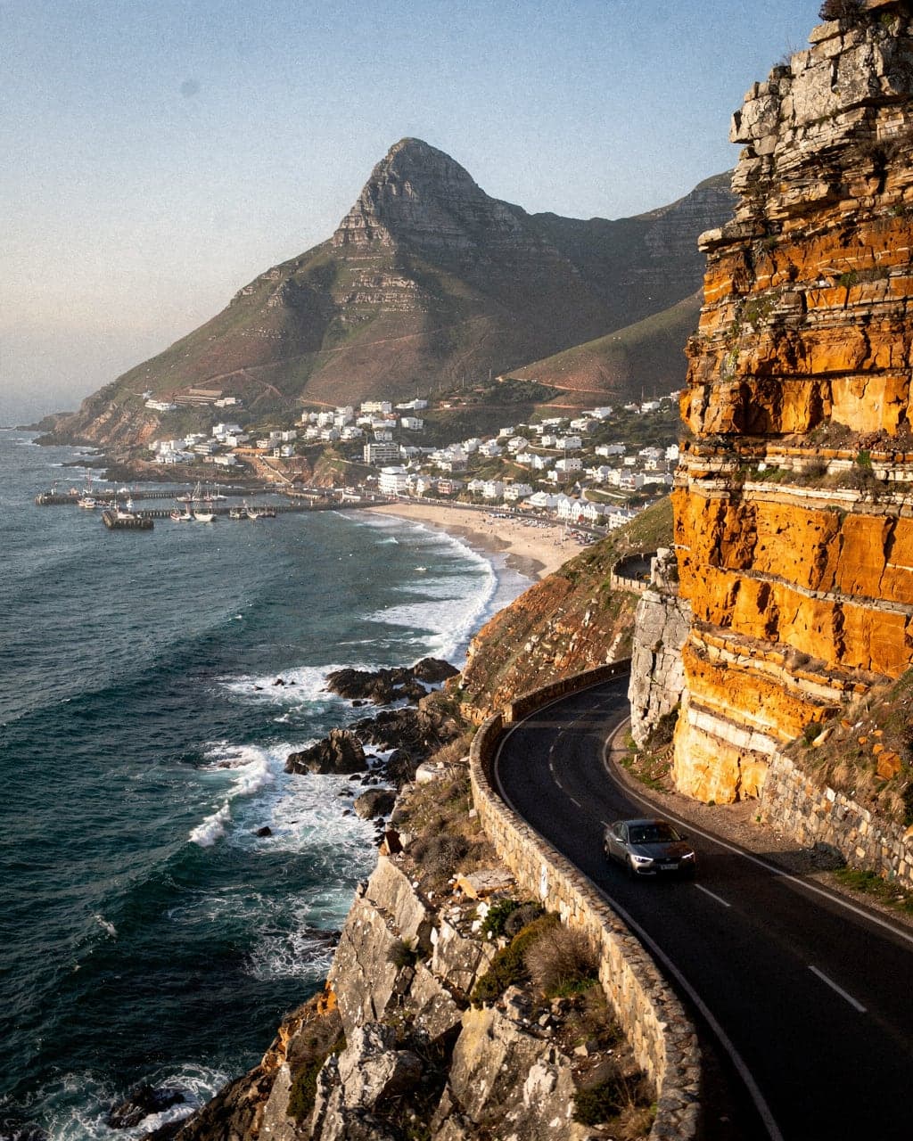 Chapman's Peak Drive cut into cliffs above Hout Bay, with the Sentinel peak and harbour below in late afternoon light
