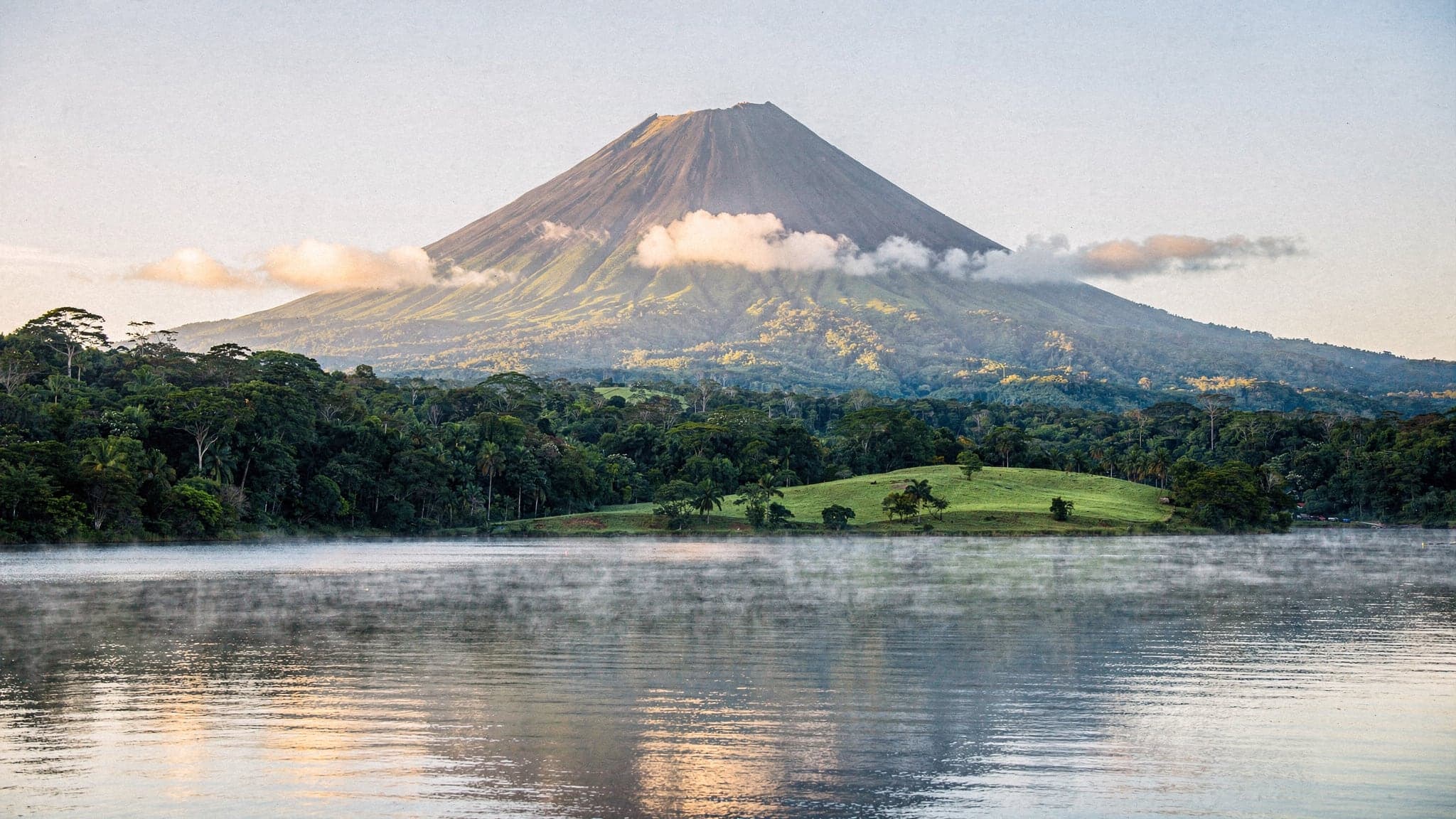 Arenal Volcano's cone rising above rainforest and Lake Arenal in clear early-morning light, with low cloud drifting over the lower slopes