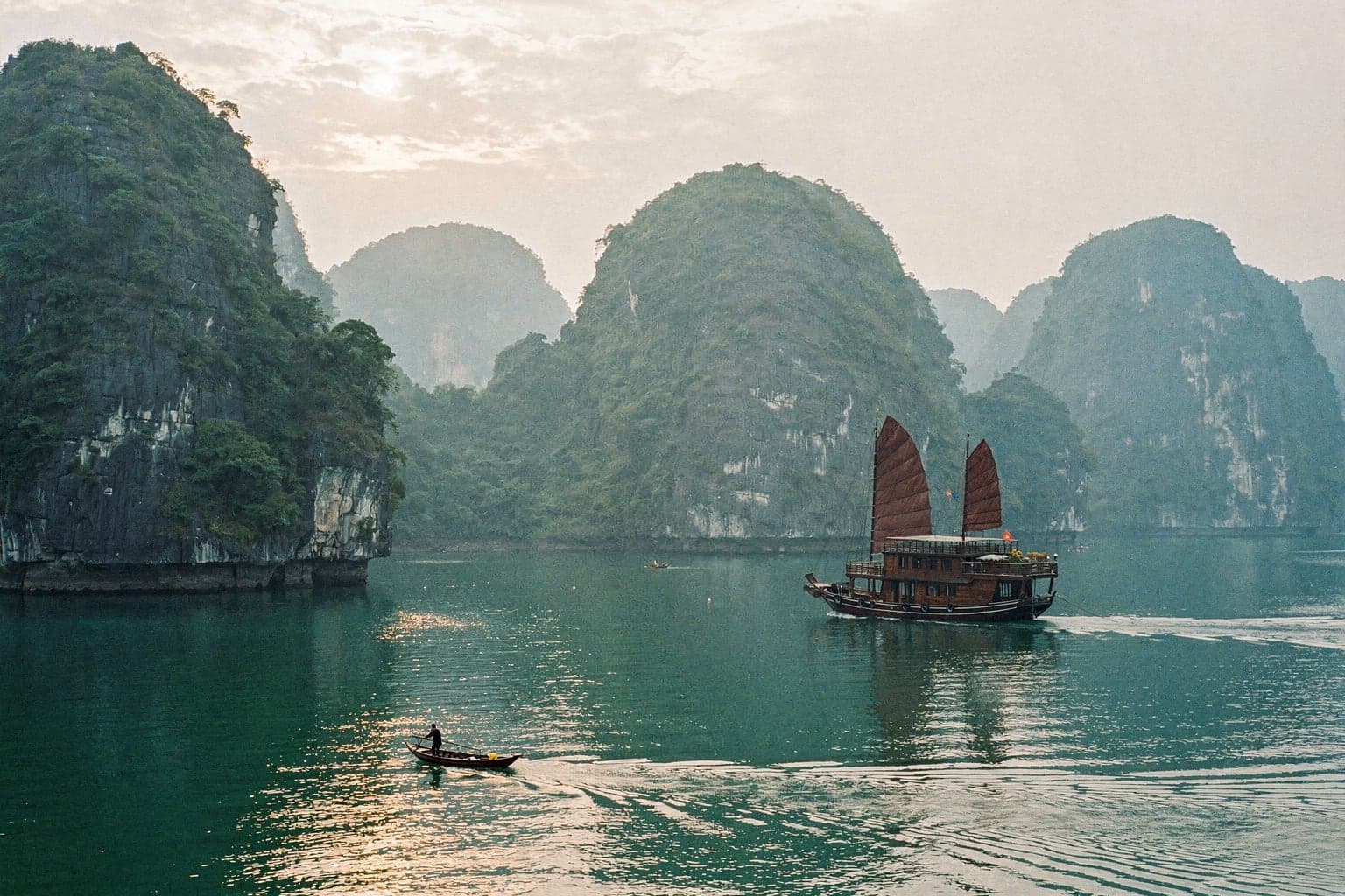 Forested limestone islands rising from calm green water in Ha Long Bay, with a wooden cruise boat at anchor