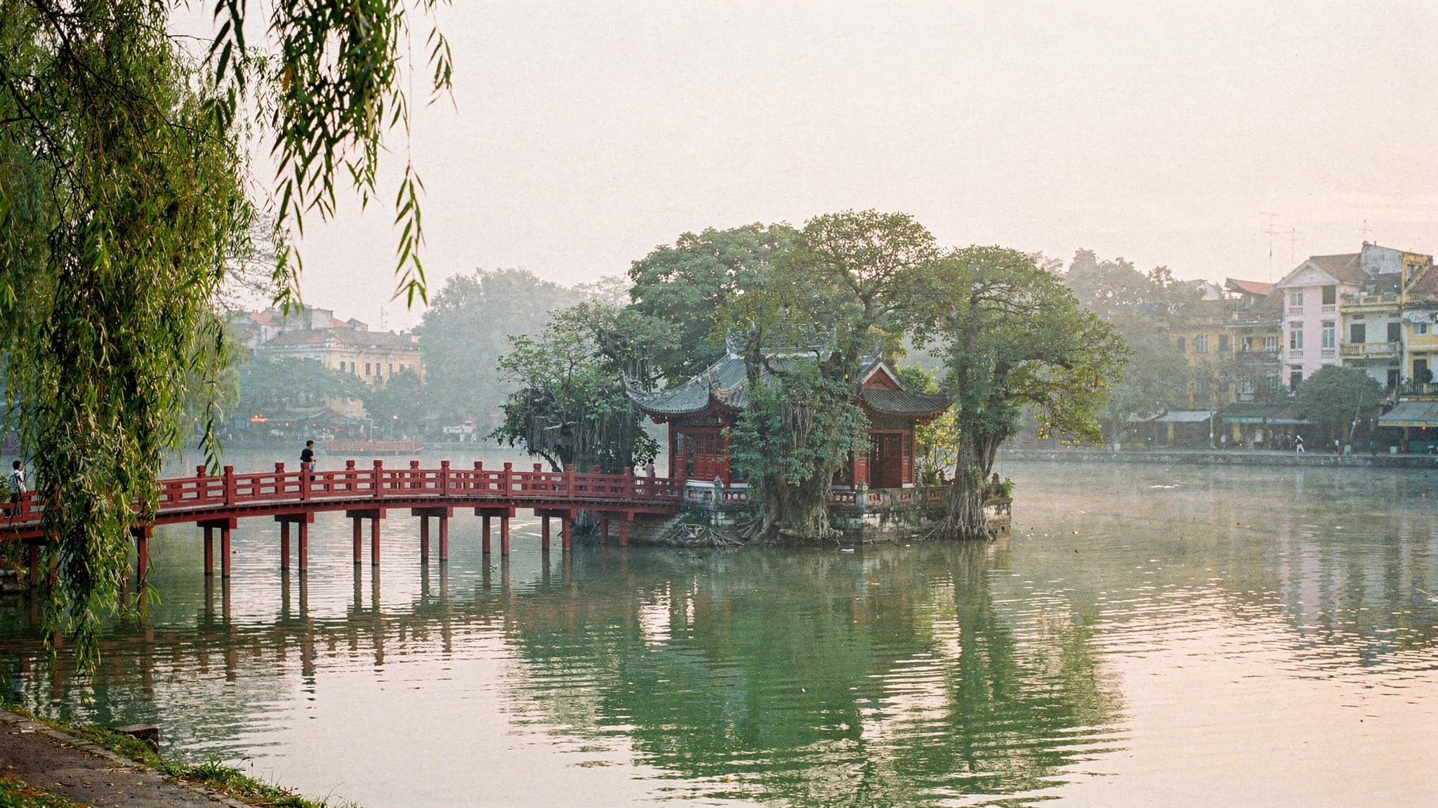 Hoan Kiem Lake at dawn, with the red Huc Bridge arching over still water to Ngoc Son Temple and early walkers on the shore