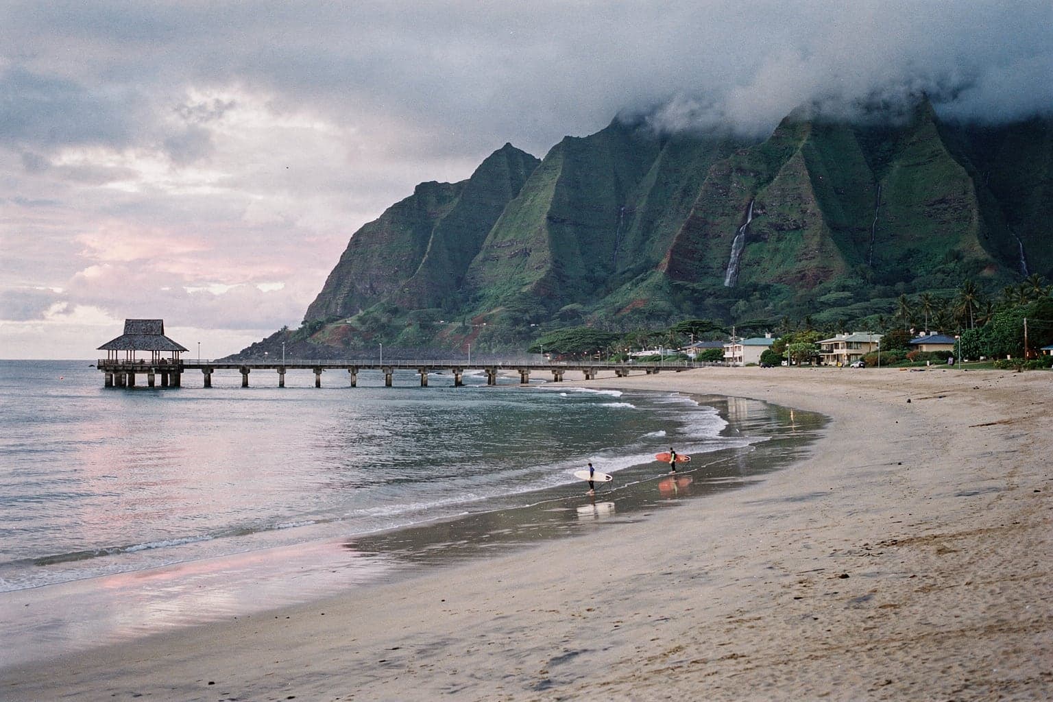 Hanalei Bay at dusk, with the long concrete pier and its roofed pavilion reaching into the bay and waterfall-streaked green mountains rising behind the town