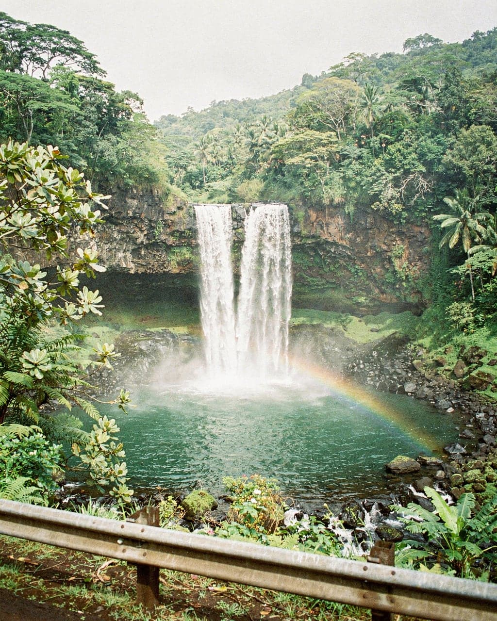 The twin plumes of Wailua Falls dropping into a green pool framed by tropical foliage, with a faint rainbow in the mist