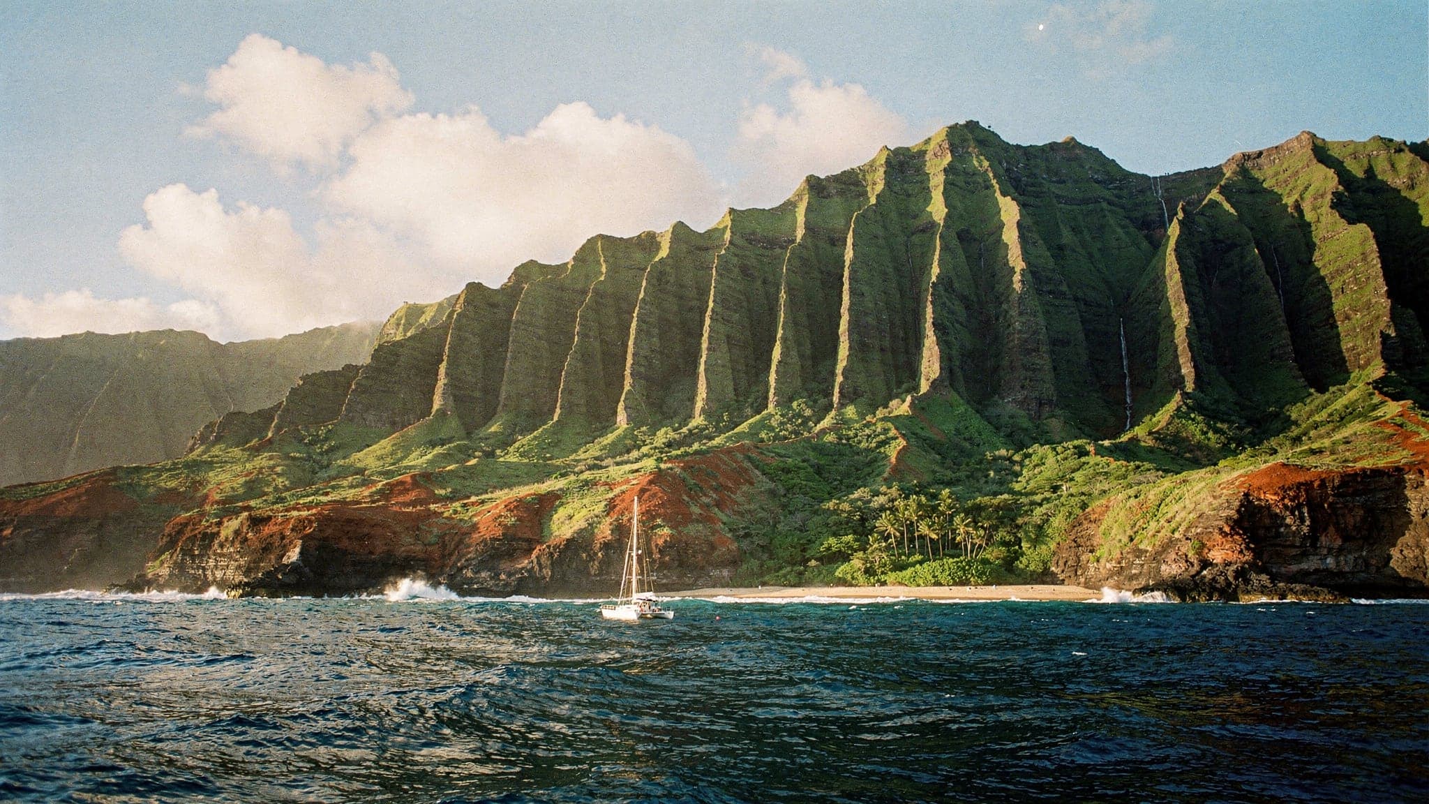 The fluted green sea cliffs of Kauai's Na Pali Coast rising from the Pacific in late-afternoon light, with a small sailing boat below them for scale