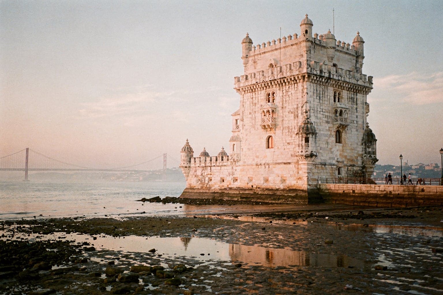 The white limestone Belém Tower at the edge of the Tagus at sunrise, with the 25 de Abril Bridge faint in the haze upriver
