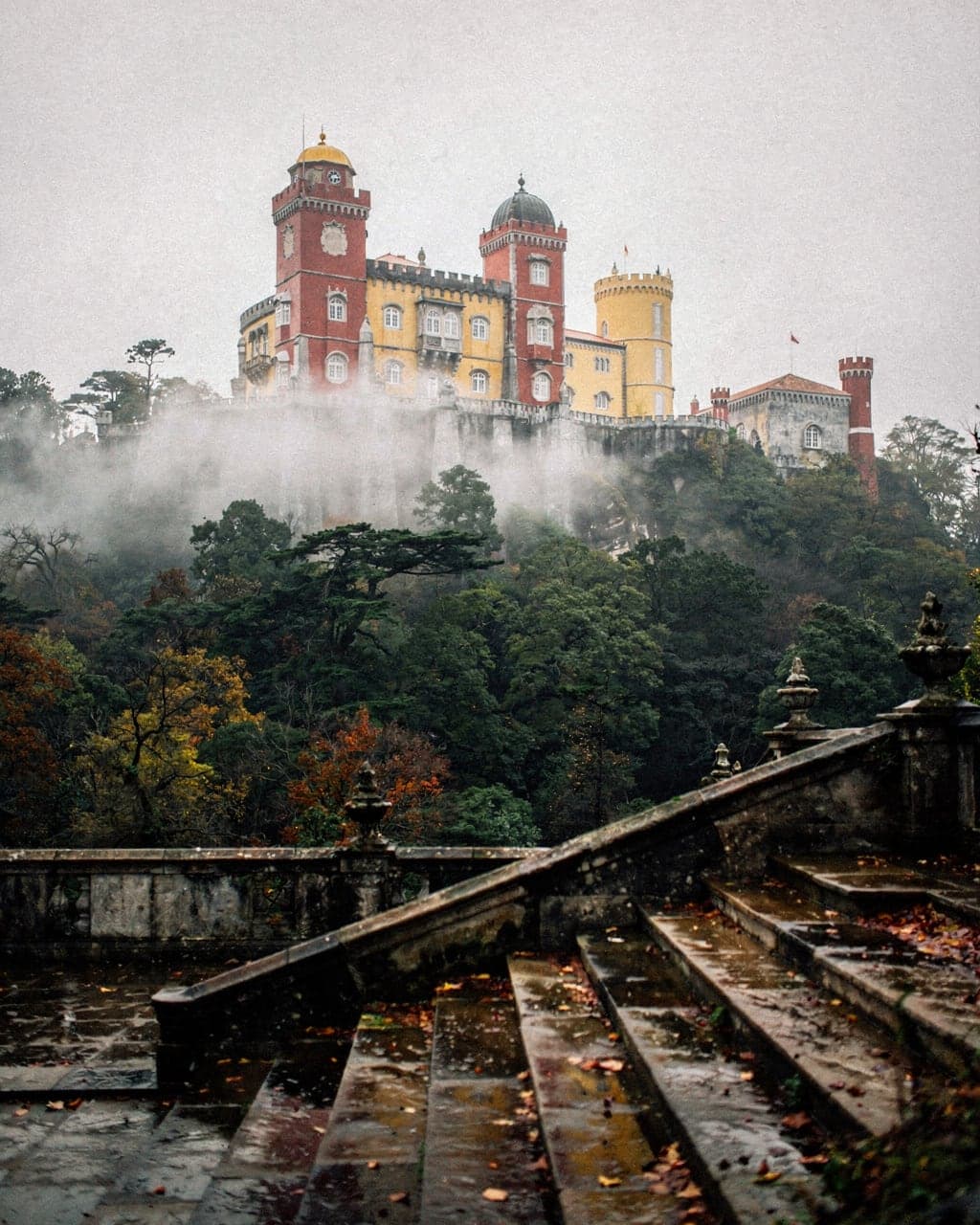 The red and yellow towers of Pena Palace rising above dark forest in morning mist, Sintra