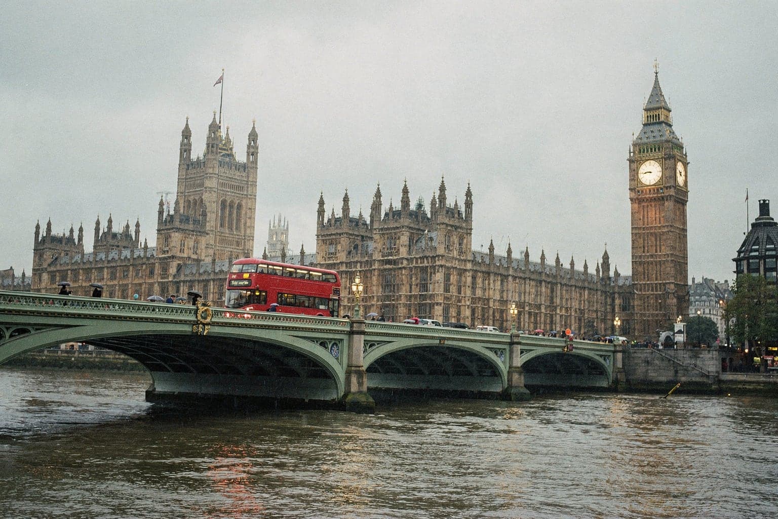 A red double-decker bus crossing Westminster Bridge in light rain, with the Elizabeth Tower behind it