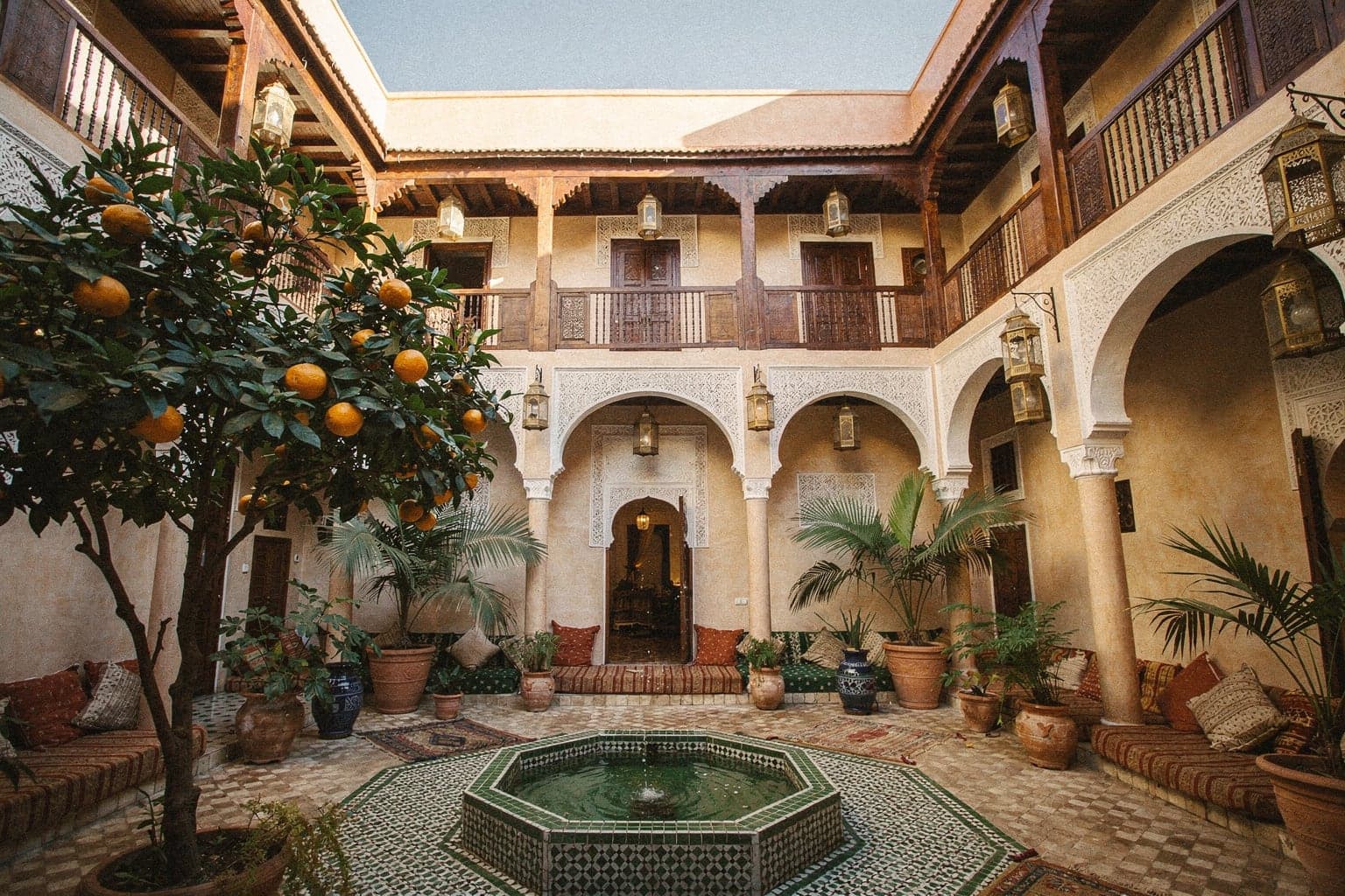 A riad courtyard with a tiled fountain, an orange tree and carved plaster arches in soft afternoon light