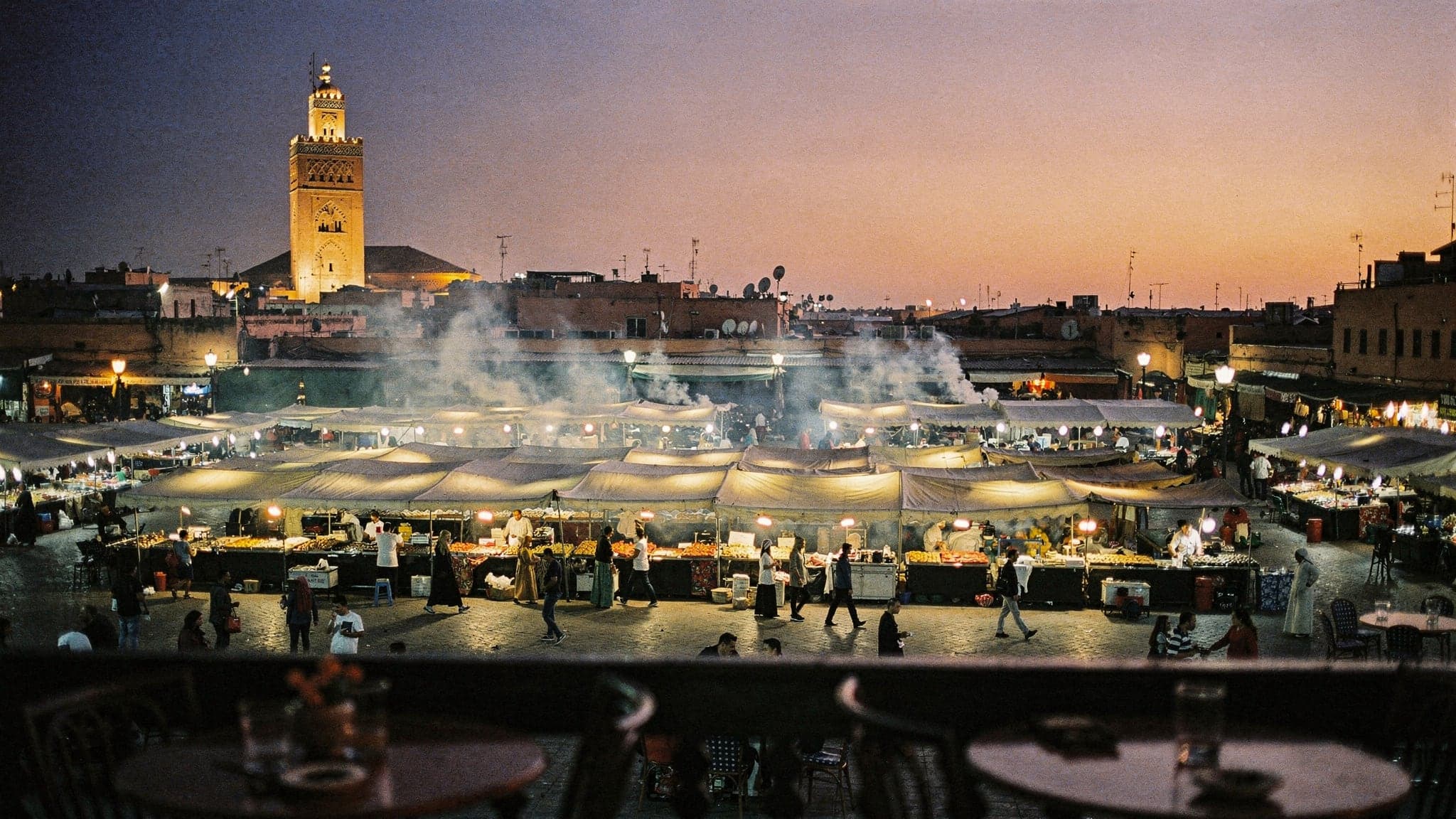 Smoke rising from food stalls on Jemaa el-Fna at dusk, with the Koutoubia Mosque's minaret against a violet sky
