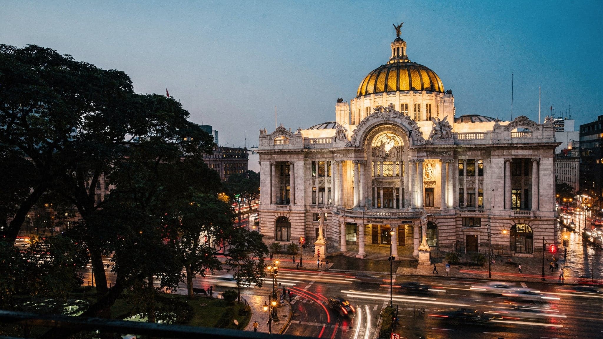 The Palacio de Bellas Artes at blue hour, its white marble facade and tiled dome floodlit above the trees of the Alameda Central and streaks of traffic