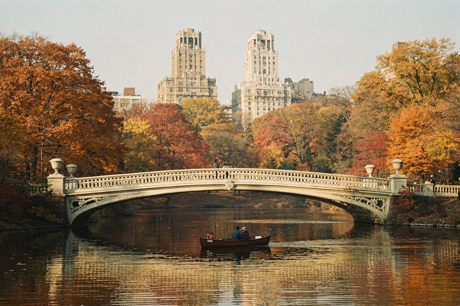 Bow Bridge in Central Park in late autumn, with orange and yellow trees reflected in the Lake and the San Remo's twin towers behind