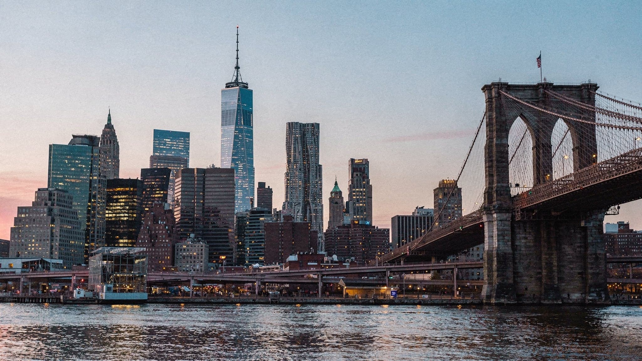 Lower Manhattan's skyline at dusk seen from Brooklyn Bridge Park, with the Brooklyn Bridge on the right and office lights coming on across the East River