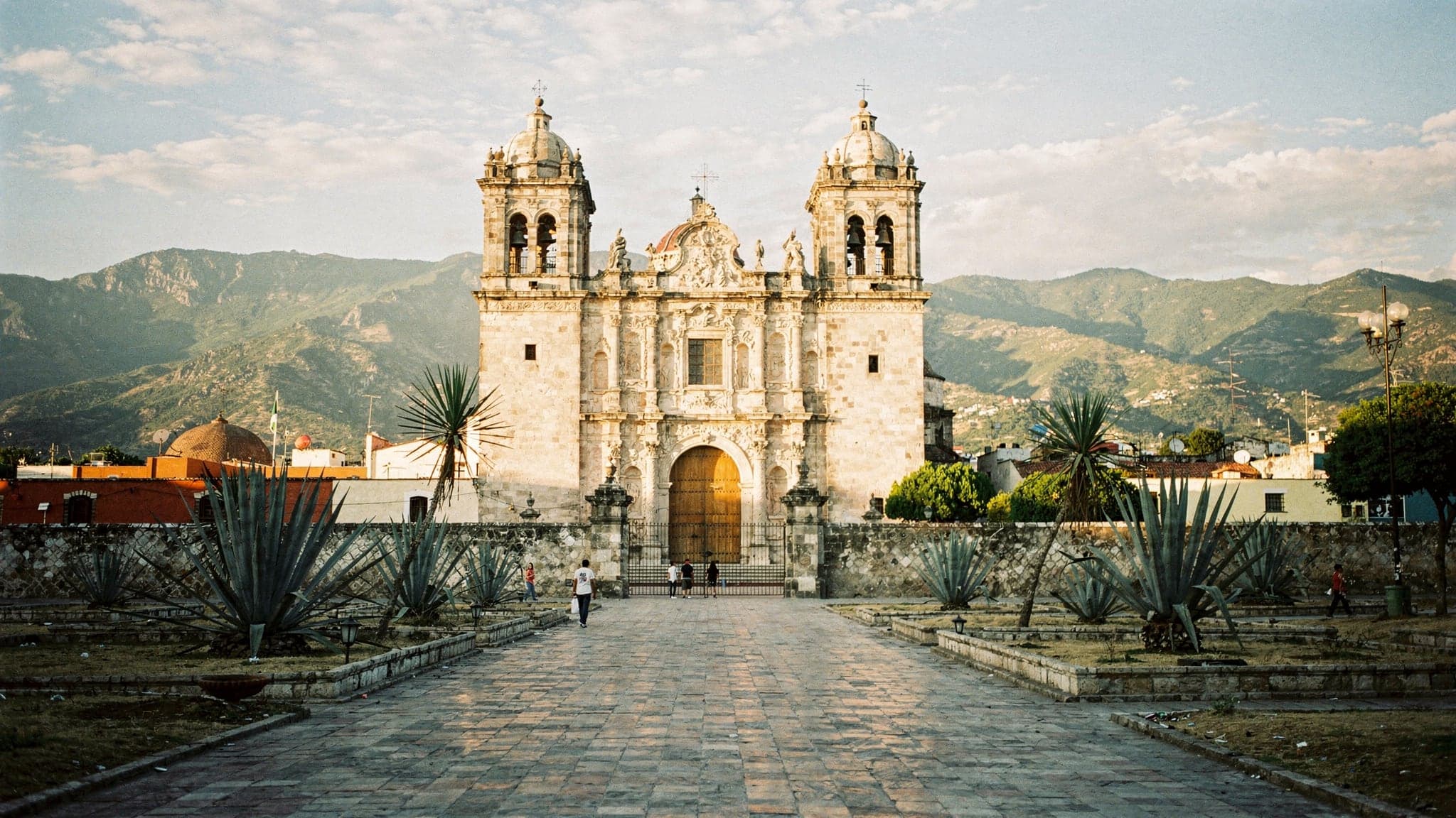 The stone facade and twin bell towers of the Templo de Santo Domingo de Guzmán in Oaxaca City at golden hour, with agave plants in the atrium garden and mountains behind