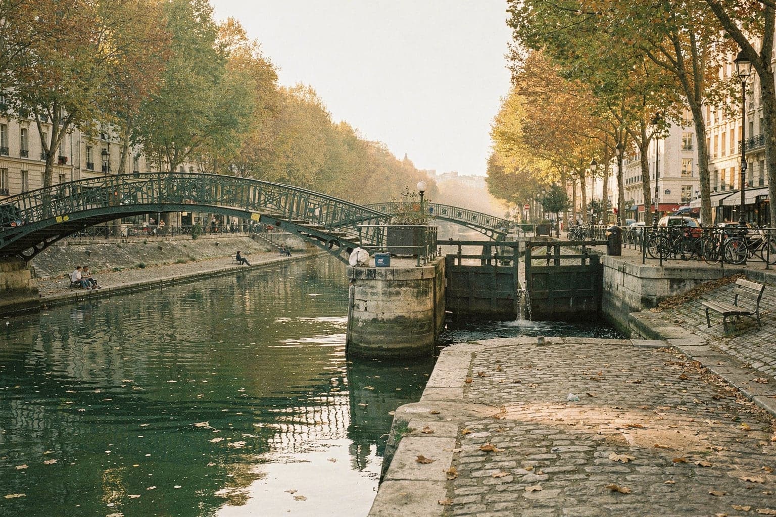 Iron footbridges over the green water of the Canal Saint-Martin at golden hour, plane trees turning amber