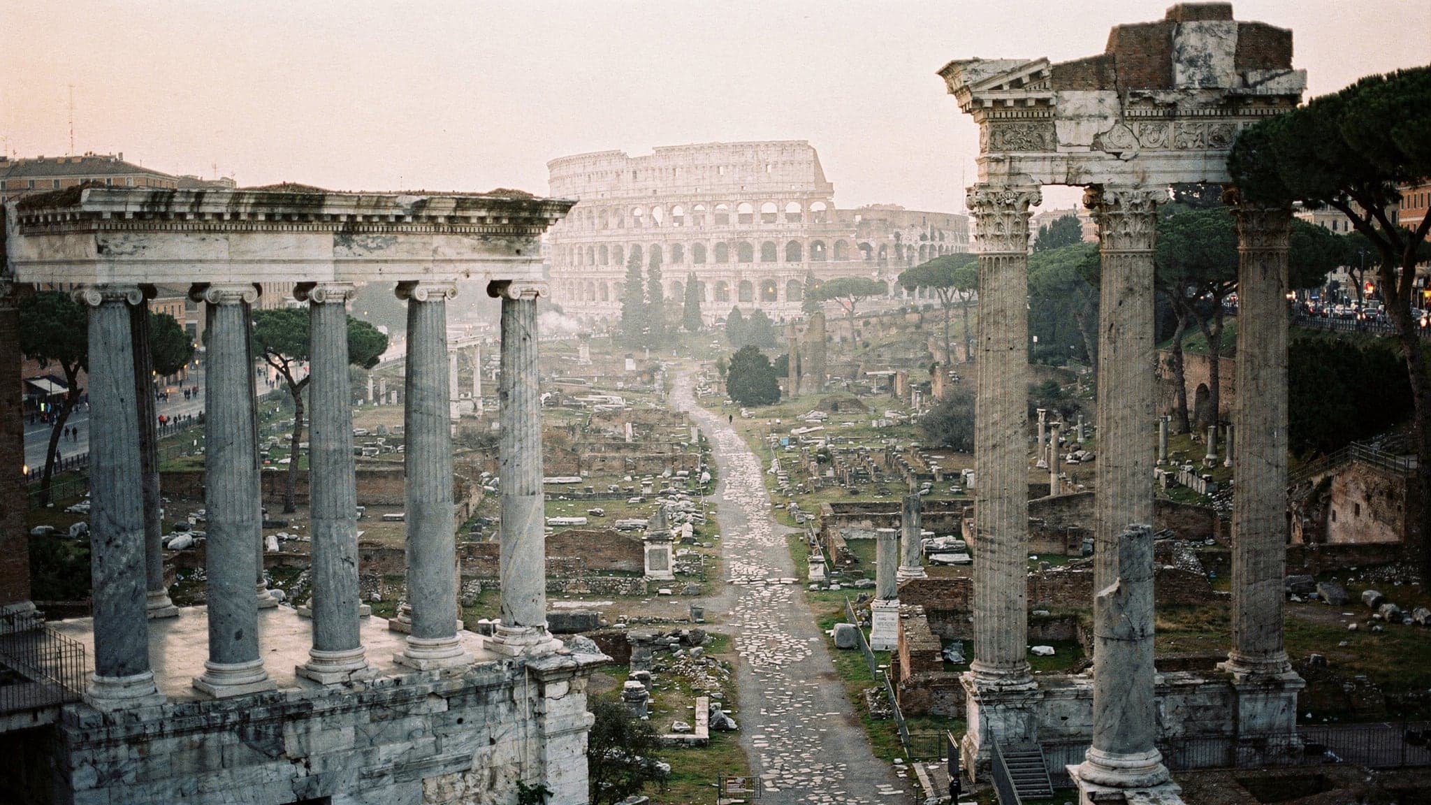 The Roman Forum at sunrise seen from the Capitoline Hill, with columns and temple ruins below and the Colosseum in the distance